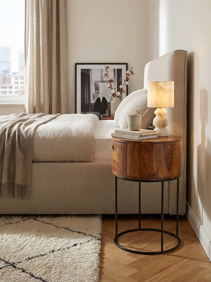 Der runde Nachttisch aus Sheeshamholz mit schwarzem Metallgestell harmoniert perfekt mit einem modernen Boxspringbett mit grauem Stoffbezug._