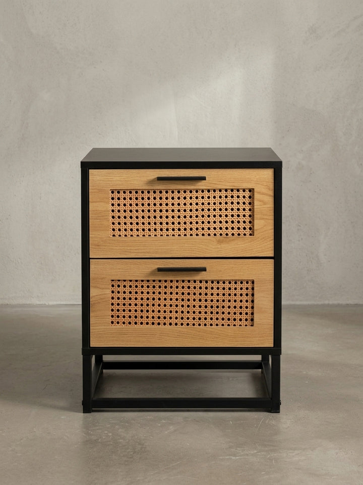 Der schwarze Nachttisch mit Rattan-Schubladen in der Dreiviertelansicht, die die Tiefe des Korpus und die filigrane Konstruktion des Eisengestells zeigt._