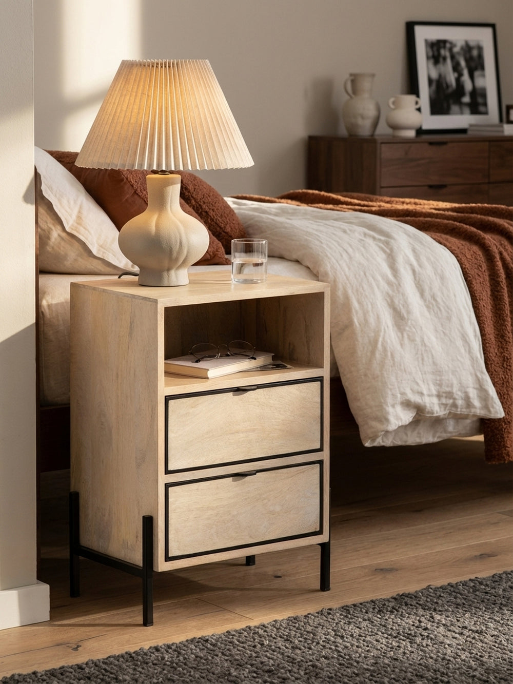 Gemütliche Abendstimmung mit dem Nachttisch aus Holz, auf dem eine Keramiklampe leuchtet und ein Buch im offenen Fach liegt._