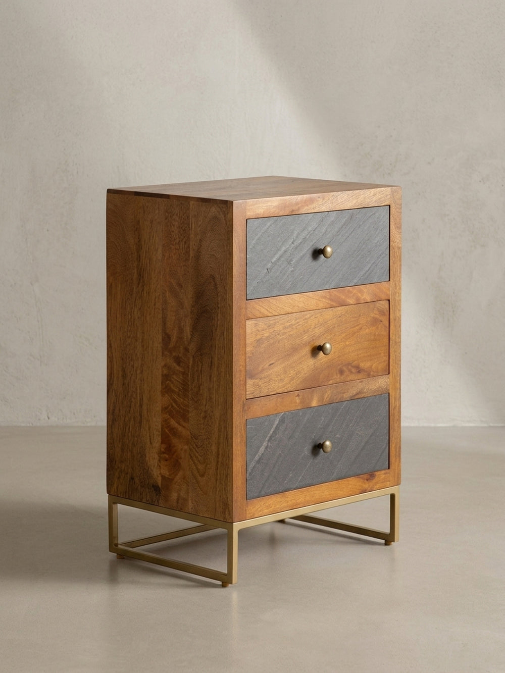 Der dreischübige Nachttisch aus braunem Mangoholz zeigt aus der 3/4-Perspektive seine schlanke Form und die Materialkombination aus Holz, Steinoptik und Metall._