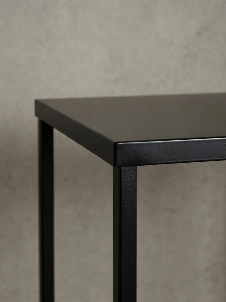 Eine alternative Perspektive auf den Kaffeetisch, die das Zusammenspiel von Licht und Schatten auf der mattschwarzen Oberfläche und dem offenen Gestell betont._