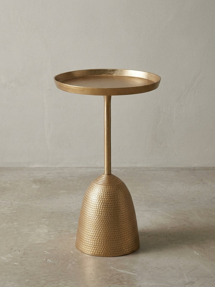 Design Beistelltisch in Gold aus einer 3/4-Perspektive, die die runde Tablett-Tischplatte und den skulpturalen, gehämmerten Standfuß aus Metall hervorhebt._