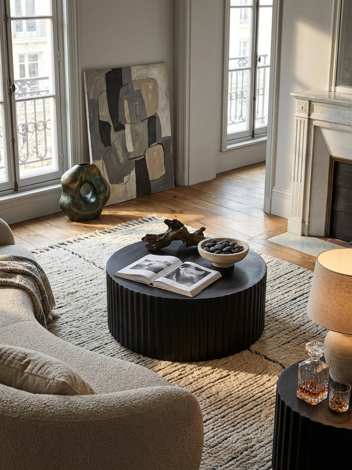 Von oben fotografierter Design-Wohnzimmertisch in Schwarz, dessen runde Form auf einem beigen Teppich vor einer grauen Couch zur Geltung kommt._