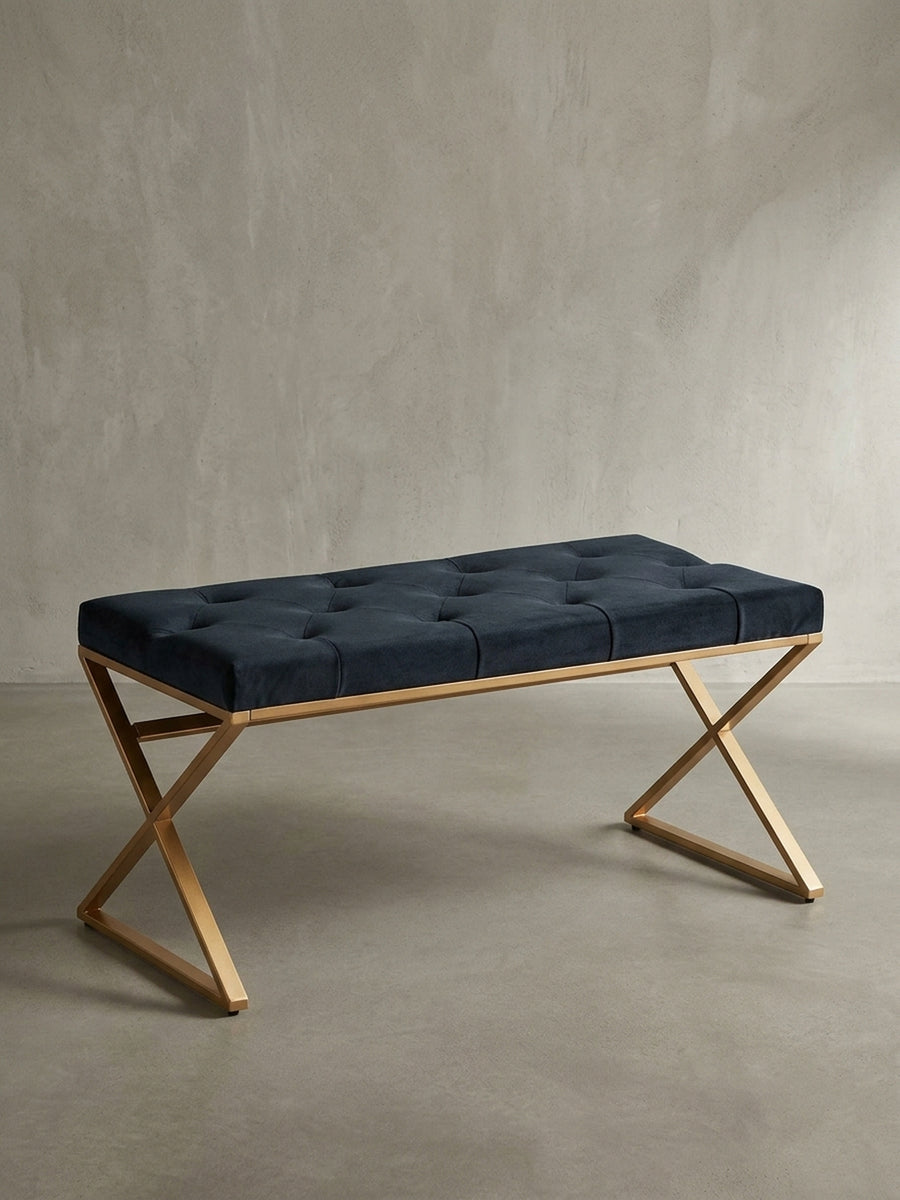 Frontalansicht der Design-Sitzbank mit schwarzem Samtpolster und gekreuztem Gestell aus goldfarbenem Edelstahl, freigestellt auf weißem Hintergrund._