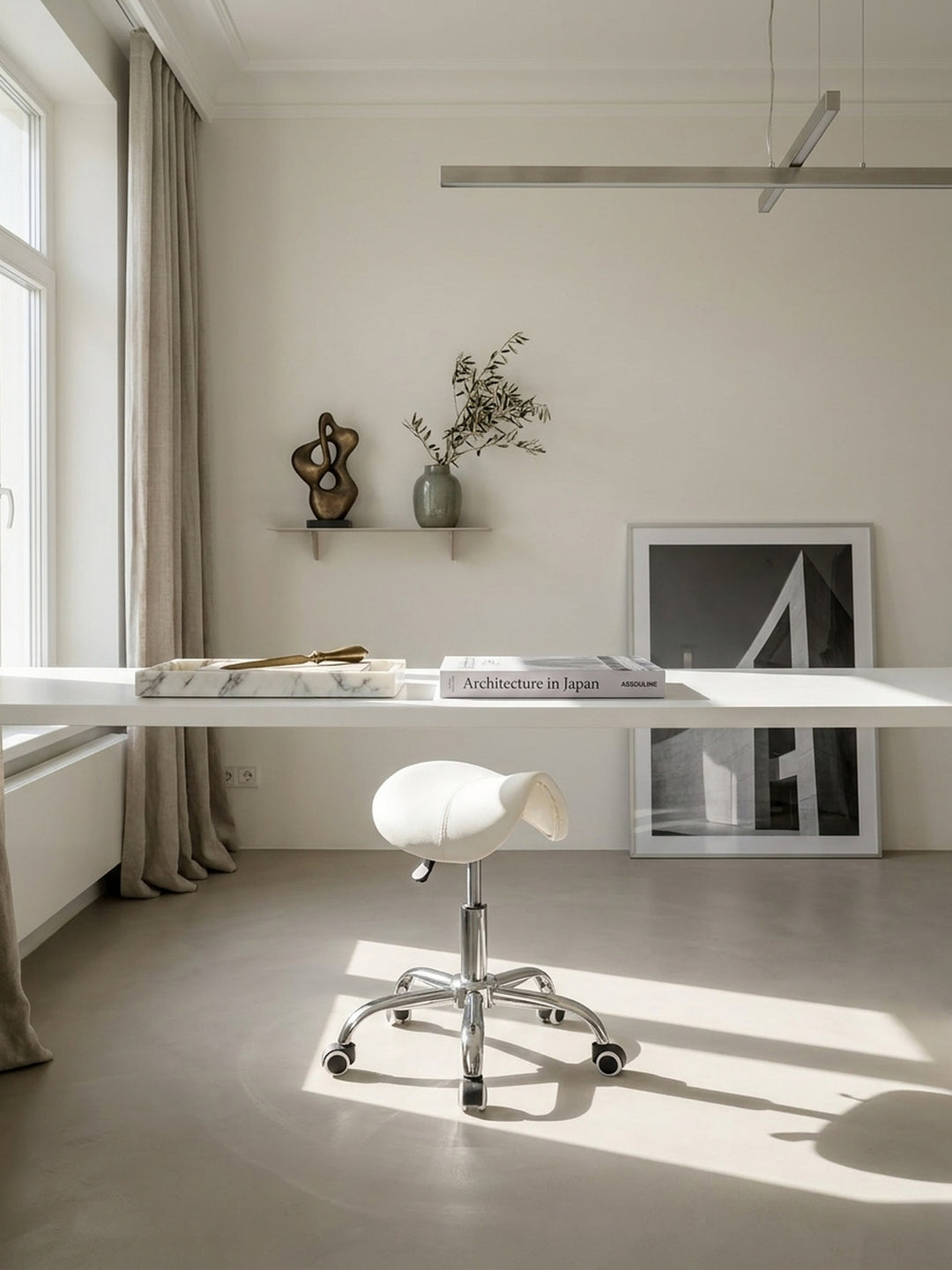 Ein weißer Sattelhocker aus Kunstleder steht vor einem schlichten Schreibtisch in einem luxuriösen, minimalistischen Home-Office mit nahtlosem, hellgrauem Mikrozementboden._