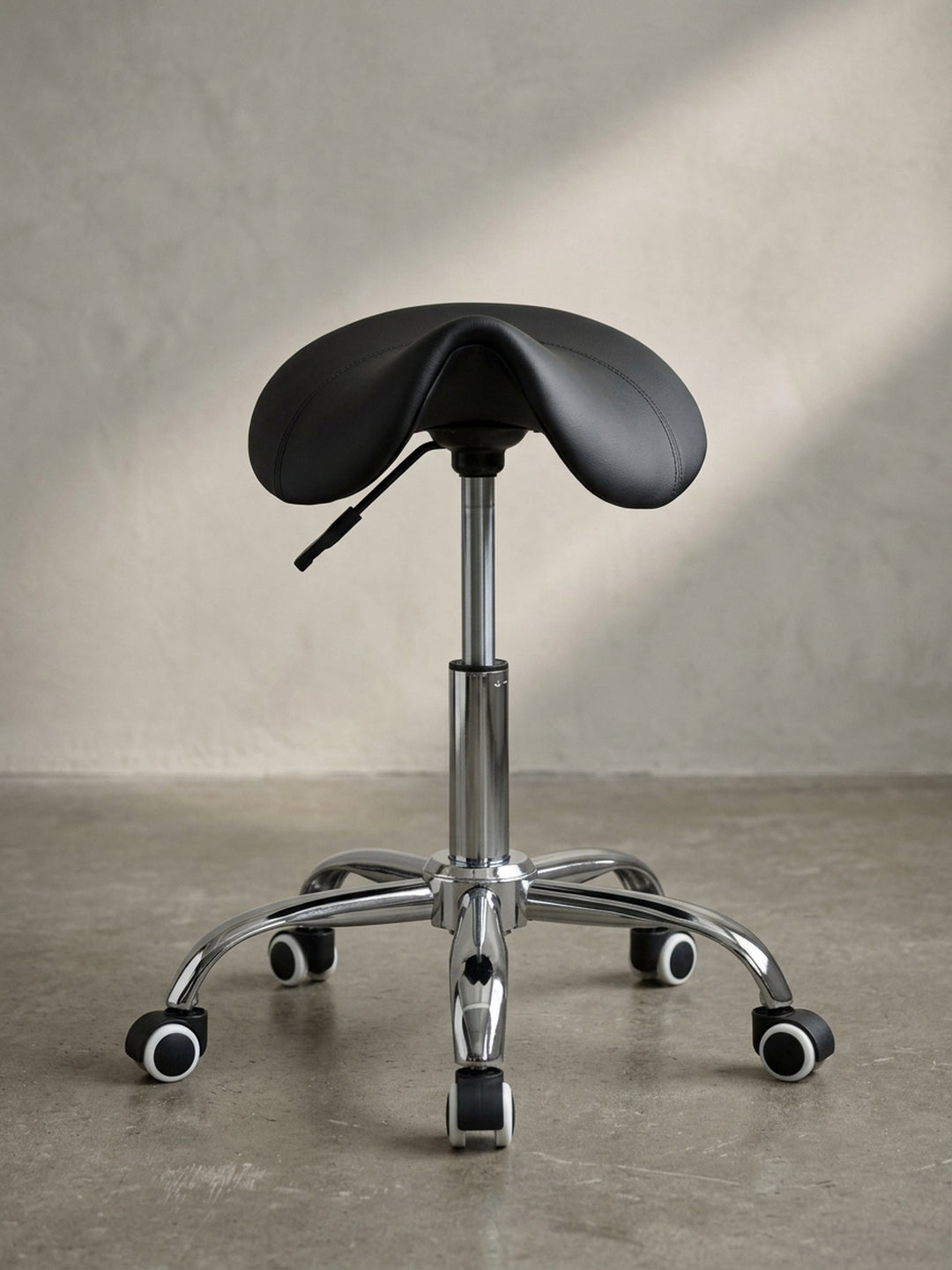 Höhenverstellbarer Rollhocker in Sattelform, Frontalansicht zeigt die ergonomische Krümmung der tiefschwarzen Sitzfläche und das glänzende Chromgestell._