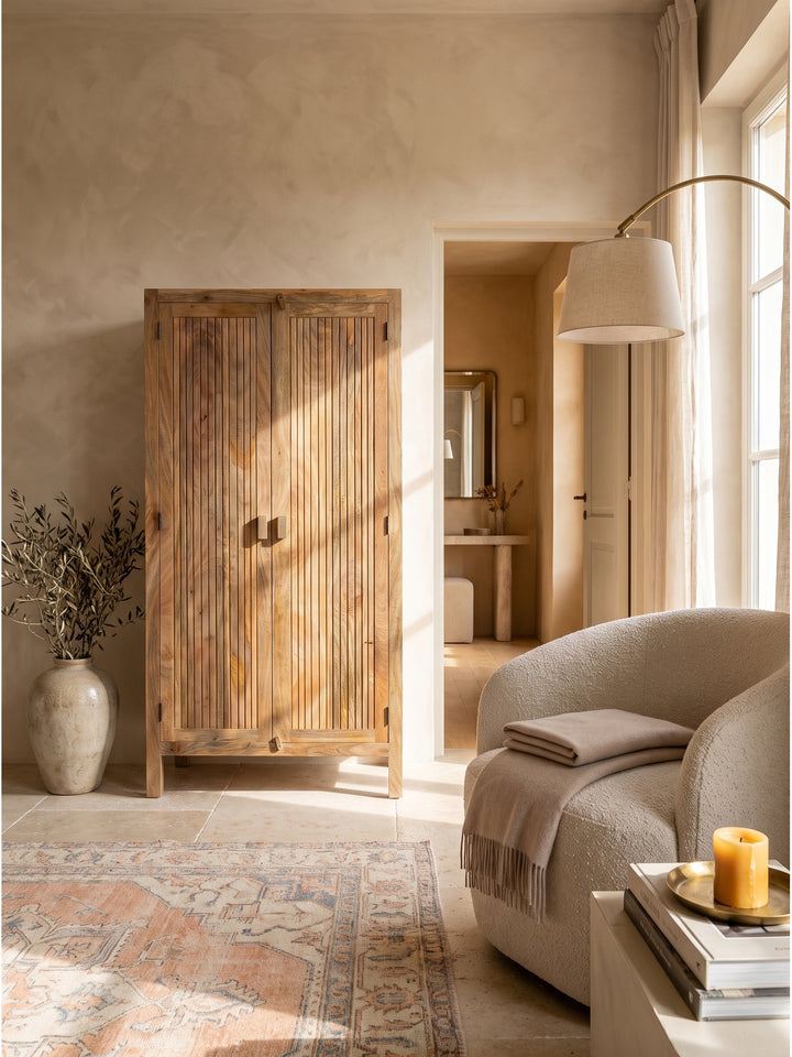 Zweitüriger Holzschrank mit Lamellendesign neben einem minimalistischen Sofa und einer skulpturalen Stehlampe vor einer Kalkputz-Wand._