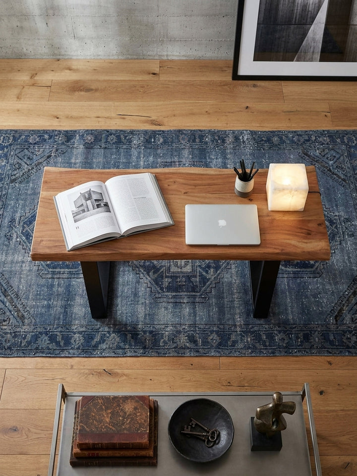 Luftaufnahme eines schmalen Computertischs aus Akazienholz mit schwarzen Metallbeinen, platziert auf einem orientalischen Teppich in einem hellen, modernen Homeoffice._