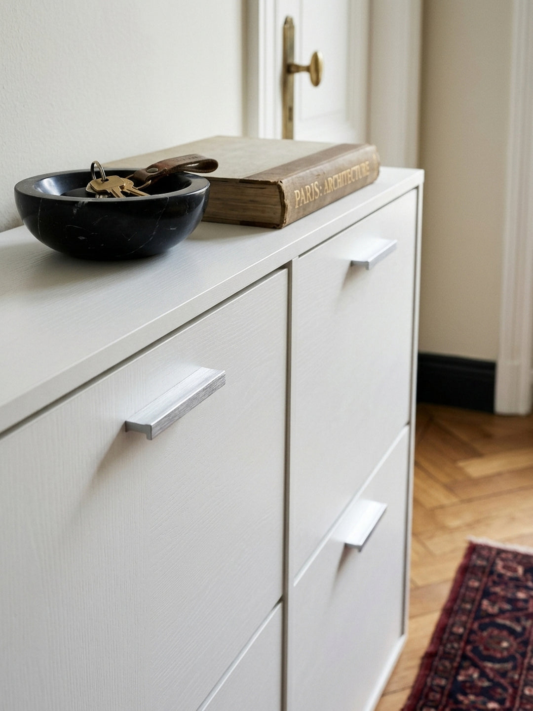 Detailaufnahme der glatten, mattweißen Oberfläche des Schuhregals. Hochwertig verarbeitete Spanplatte trifft auf elegante, gebürstete Aluminiumgriffe. Flurschrank Holz in reinem Weiß._