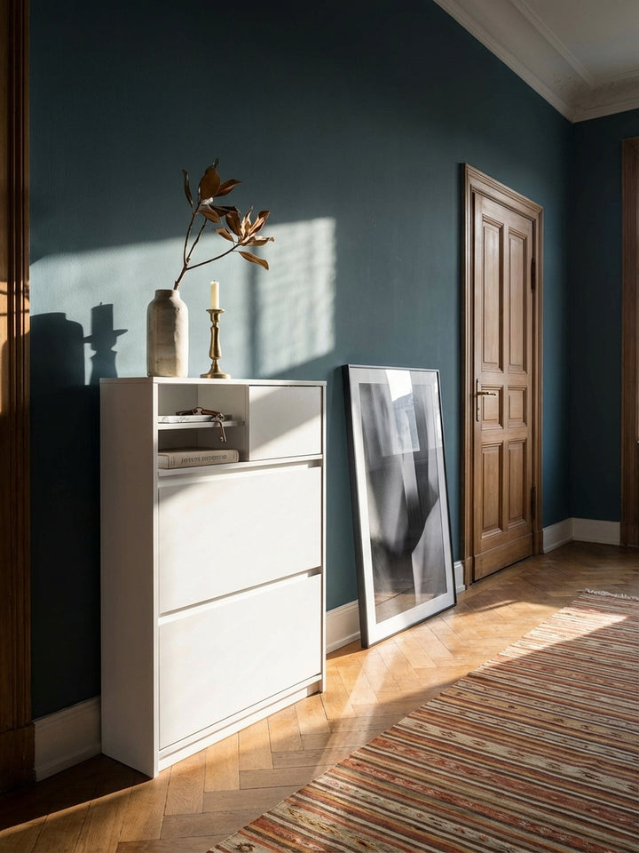 Moderner weißer Schuhschrank in einem eleganten Altbau-Flur mit dunkler Wandfarbe und Fischgrätparkett, dekoriert mit einer Vase und einem Bild._