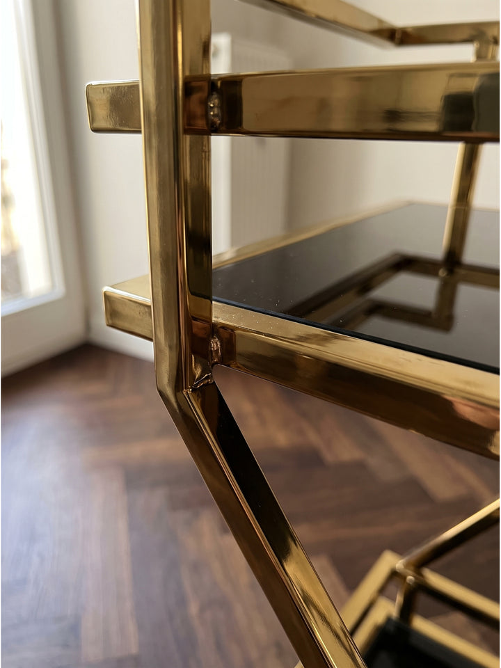 Nahaufnahme der schwarzen Glasablage und des polierten goldenen Stahlrahmens des Getränkewagens mit sichtbaren Schweißnähten._