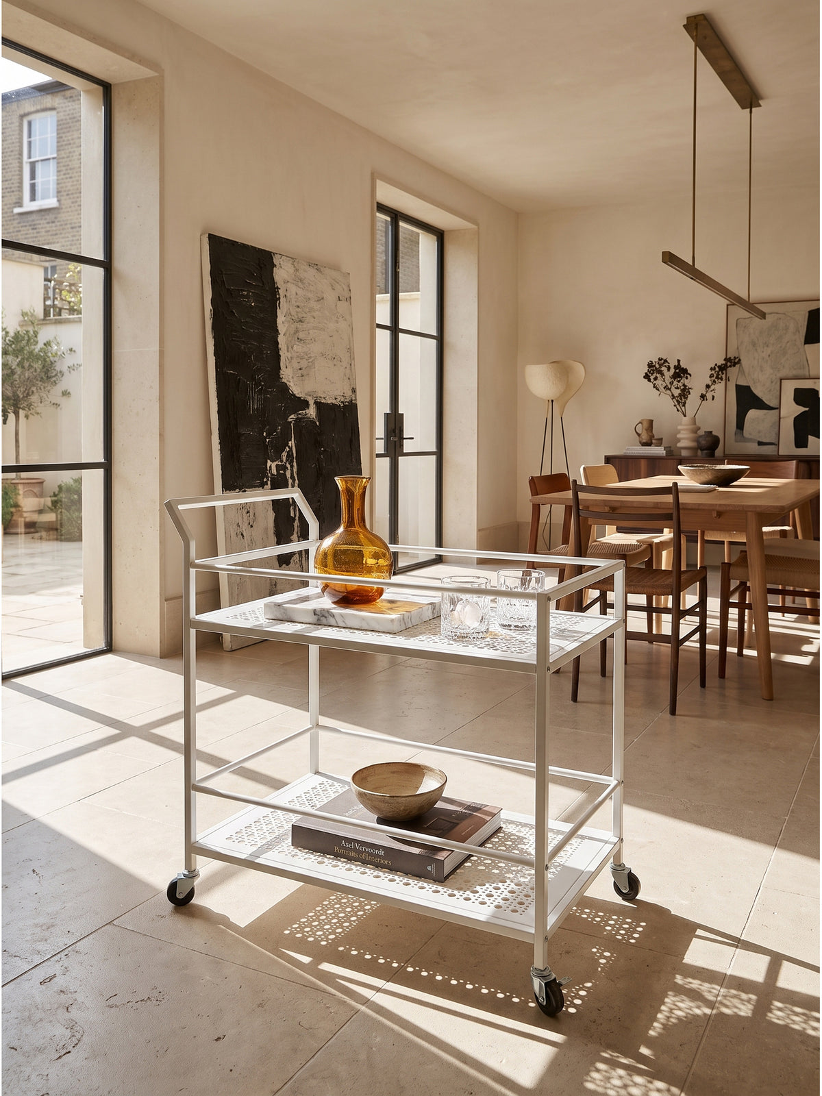 Minimalistischer Servierwagen aus weißem Metall im luxuriösen Esszimmer arrangiert mit bernsteinfarbenem Glas auf hellem Kalksteinboden_