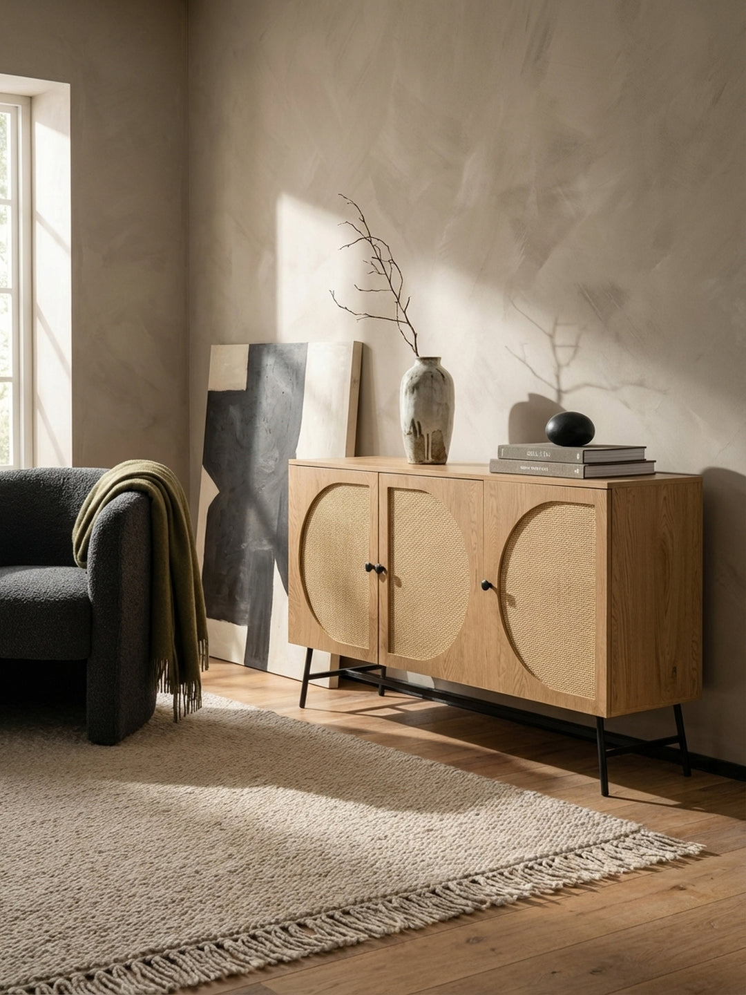 Das Rattan-Sideboard kombiniert mit einem Sessel aus dunklem Bouclé-Stoff, was einen eleganten Materialmix im modernen Wohnbereich erzeugt._