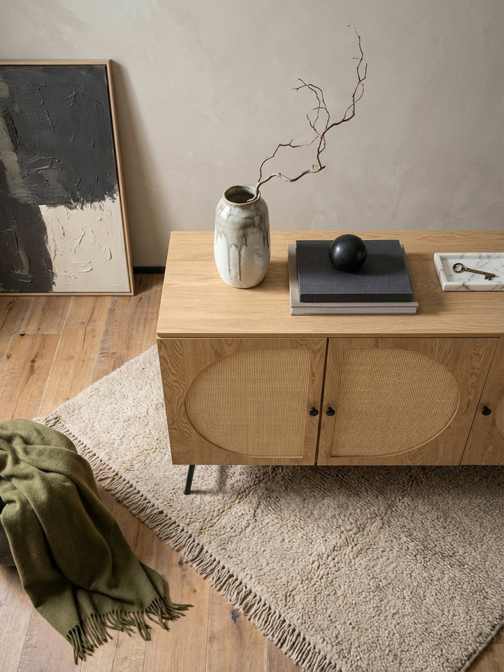 Draufsicht auf ein Wohnzimmer mit der Kommode aus Eichenholz und Rattan, die als Mittelpunkt neben einem grauen Sofa auf einem beigen Wollteppich steht._