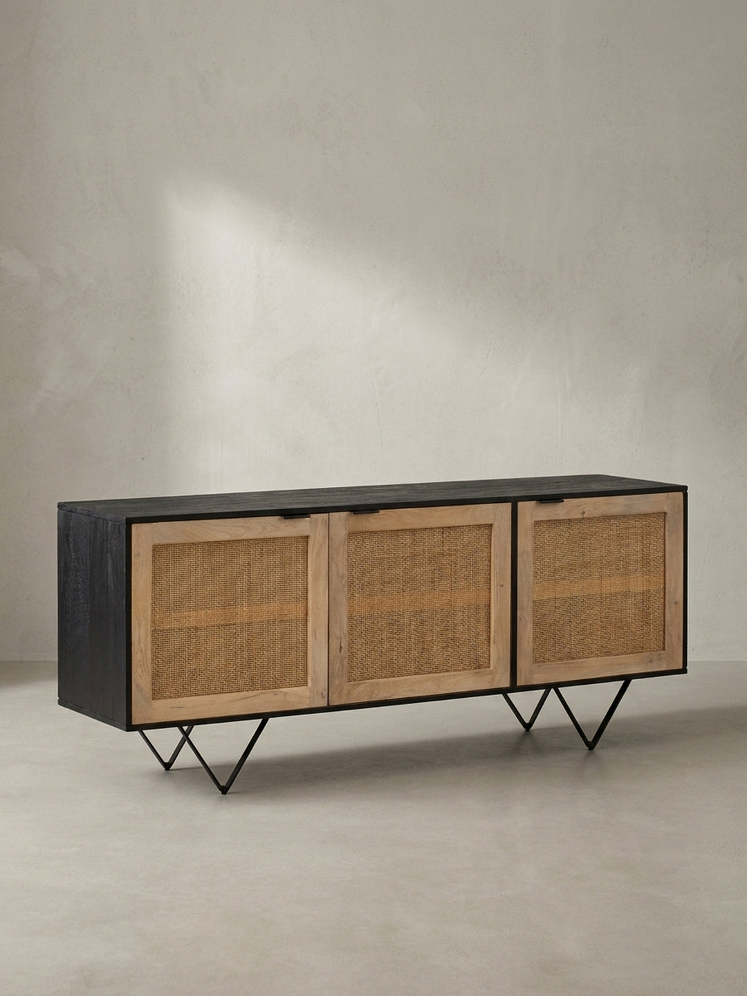 Angewinkelte Darstellung der Massivholz-Kommode mit geflochtenen Rattanfronten und schwarzen V-förmigen Metallbeinen auf weißem Grund._