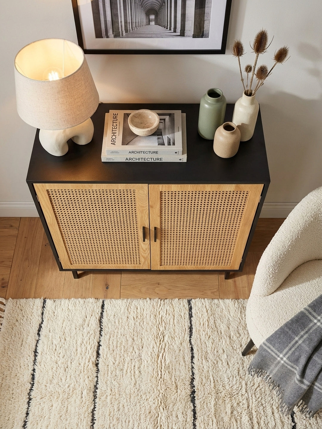 Draufsicht auf eine elegante Kommode in Schwarz und Rattan, arrangiert mit einer Tischlampe und Büchern auf einem cremefarbenen Wollteppich._