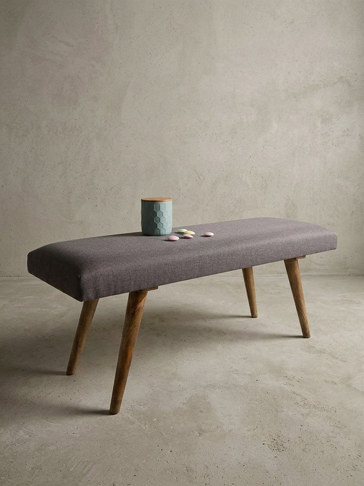 Frontale Ansicht der 117 cm breiten Sitzbank in Grau. Die klare, rechteckige Silhouette der Polsterbank im Retro-Stil wird durch vier ausgestellte Beine aus lackiertem Mangoholz getragen._