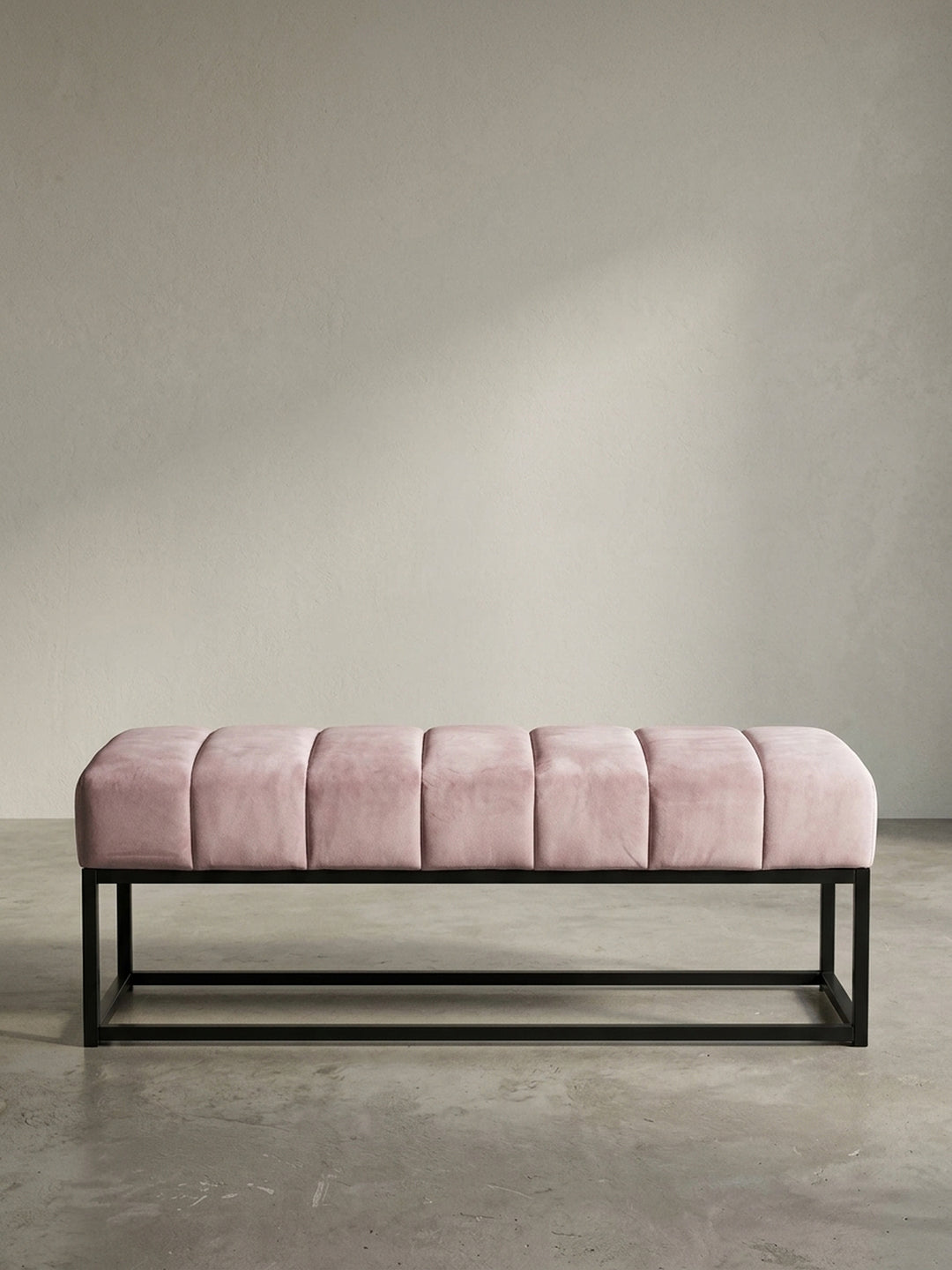 Schräge 3/4-Ansicht der rosa Samt-Polsterbank, die die Tiefe der gesteppten Sitzfläche und die klare Linienführung des Gestells zeigt._