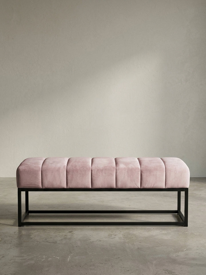 Schräge 3/4-Ansicht der rosa Samt-Polsterbank, die die Tiefe der gesteppten Sitzfläche und die klare Linienführung des Gestells zeigt._