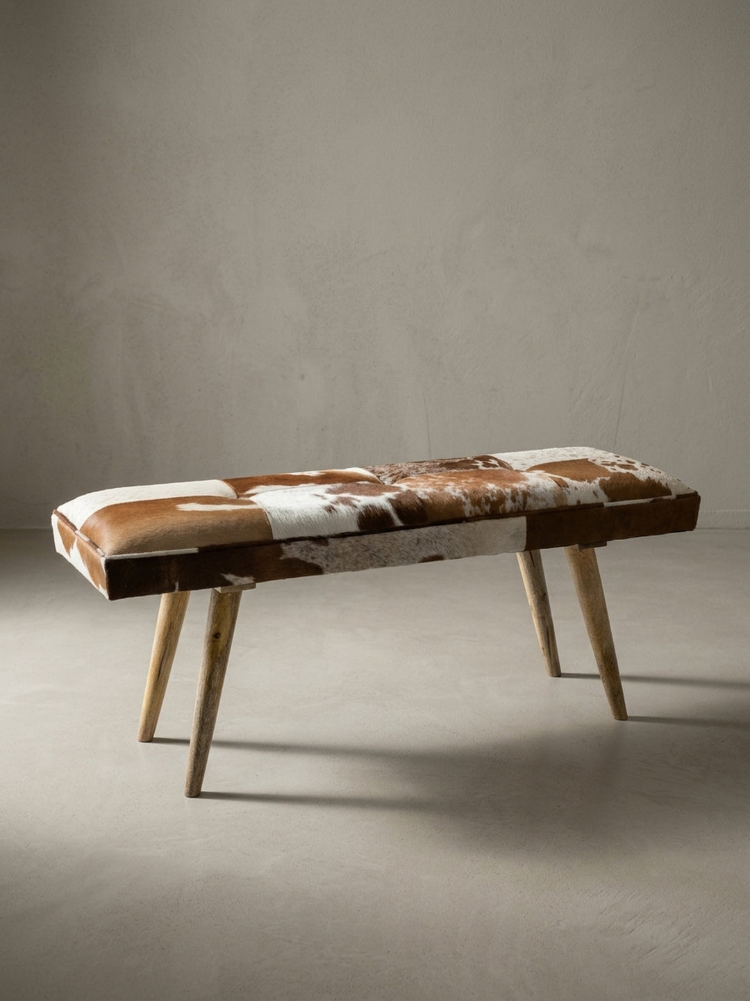 Frontale Ansicht der 120 cm breiten Sitzbank im Country Western Style. Die gepolsterte Sitzfläche aus Ziegenfell und die schrägen Massivholzbeine aus Mango bilden ein Unikat._