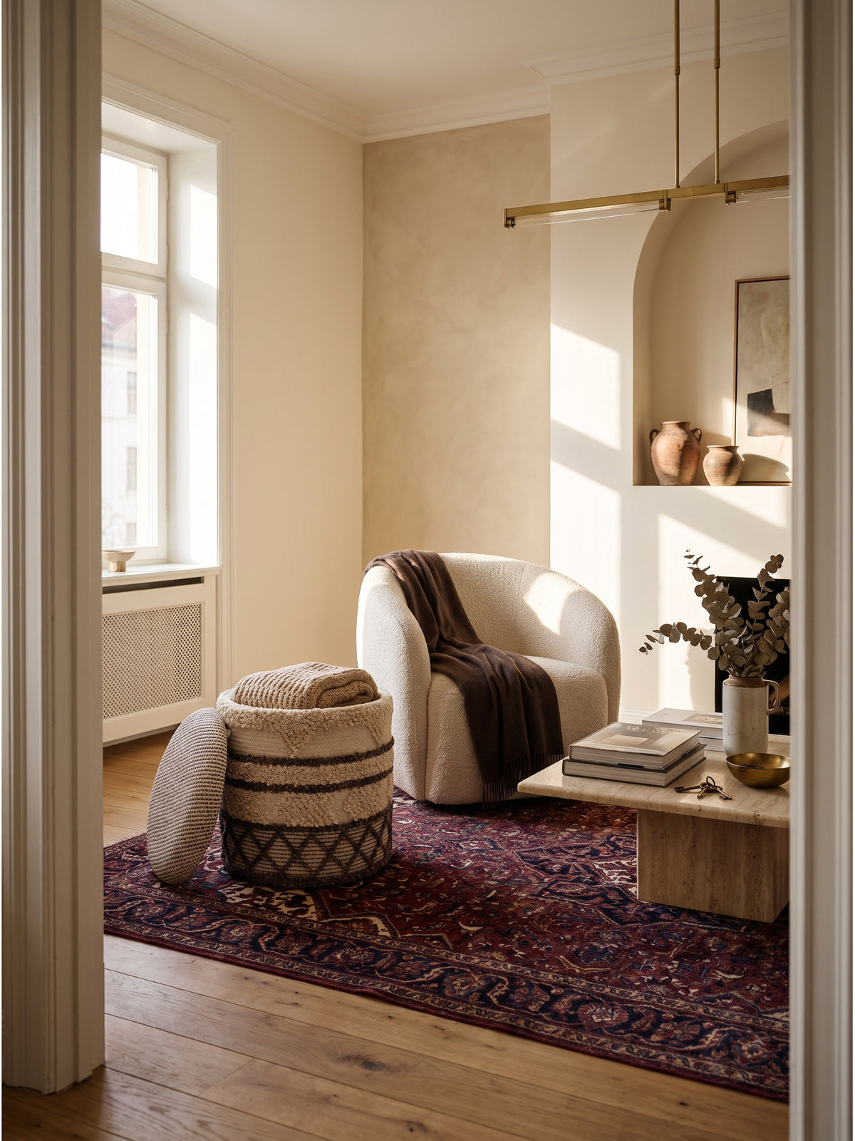 Runder Sitzhocker aus beige-brauner Wolle mit handgewebtem Boho-Muster in einem organisch modernen Wohnzimmer vor sandfarbener Wand._