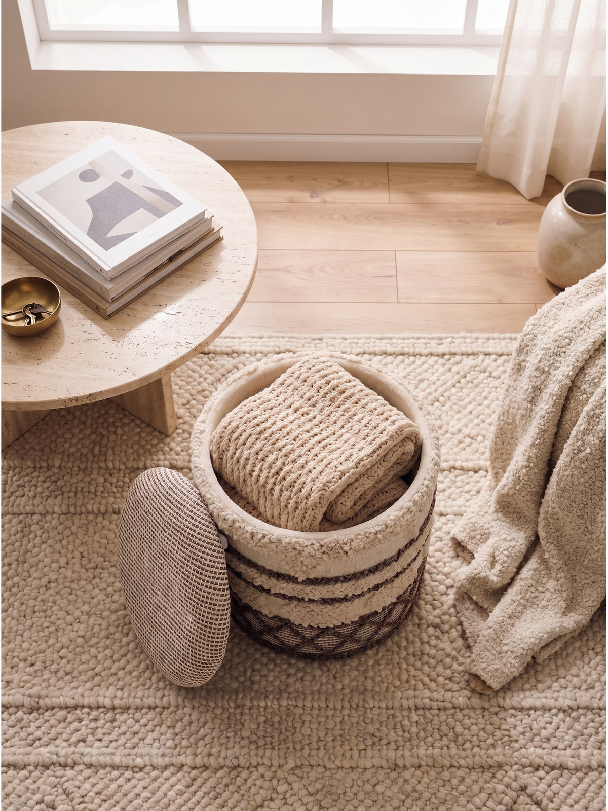 Blick von oben auf einen zylindrischen Stoffhocker mit geometrischem Webmuster neben einem cremefarbenen Bouclé-Sessel und Travertin-Tisch.