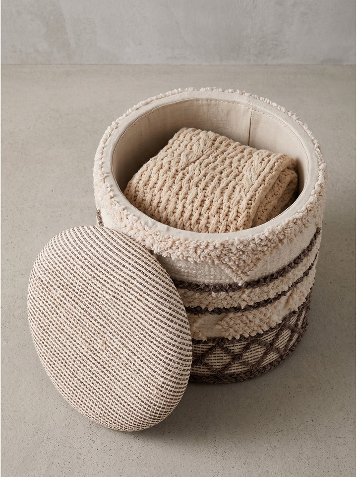 Halbseitige Ansicht des textilen Stauraumhockers mit sichtbarem Innenfach und aufgelegter Kuscheldecke in einer hellen Studioumgebung._