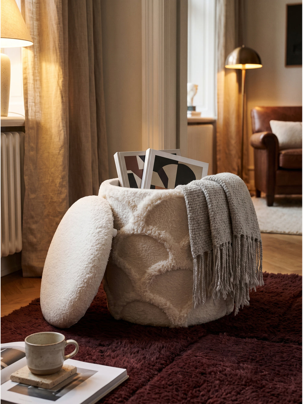 Runder Stauraumhocker mit Zeitschriften und Strickdecke dekoriert vor einem bequemen Loungesessel aus Bouclé_