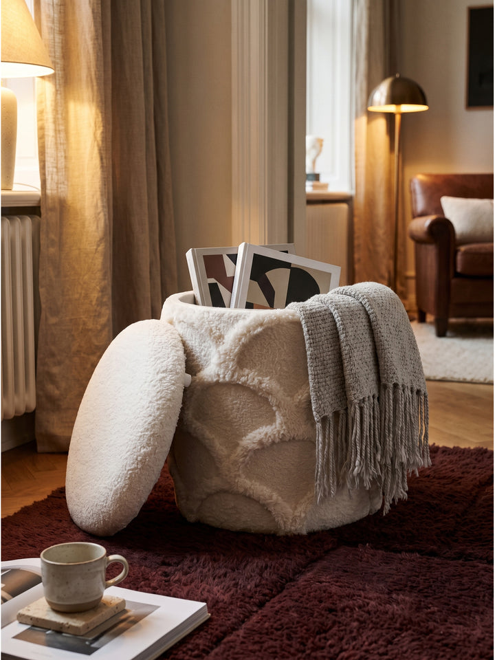 Runder Stauraumhocker mit Zeitschriften und Strickdecke dekoriert vor einem bequemen Loungesessel aus Bouclé_