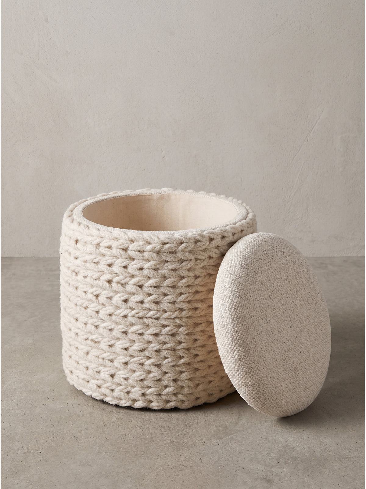 Als gemütliche Sitzgelegenheit präsentiert sich der cremeweiße Stauraum-Hocker mit seiner texturierten Hülle aus grobmaschiger Wolle._