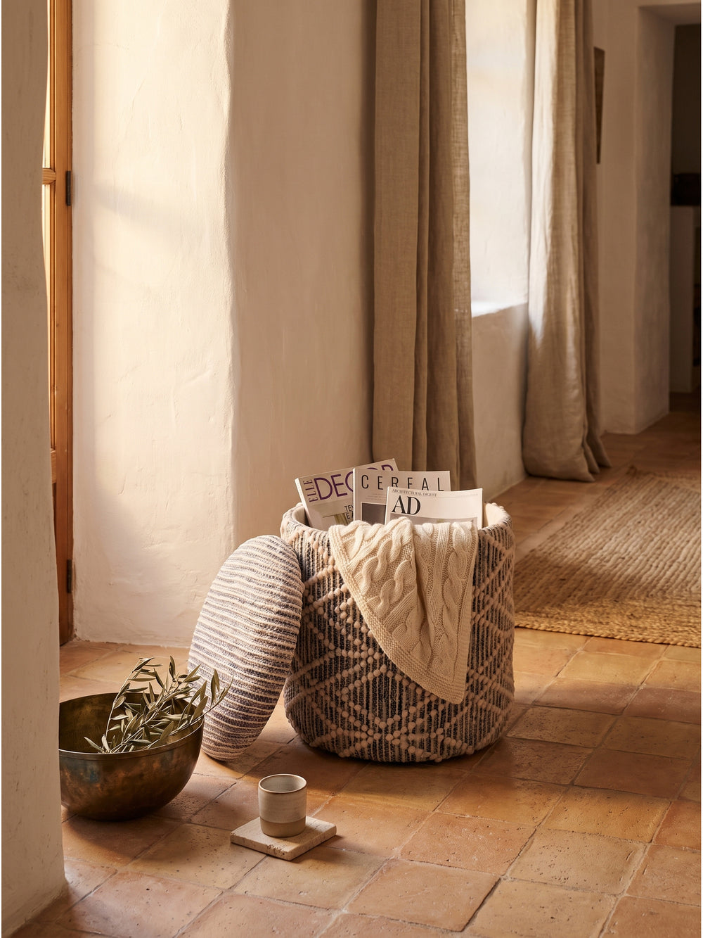 Nahaufnahme des grauen Poufs mit einem grob gestrickten Plaid und Zeitschriften im Stauraum auf einem wolligen Schlingenteppich_