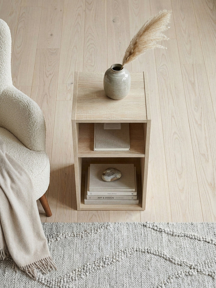 Draufsicht auf eine gemütliche Leseecke mit einem kleinen Nischenregal aus beschichtetem Holzwerkstoff neben einem Sessel auf einem handgewebten grauen Wollteppich._