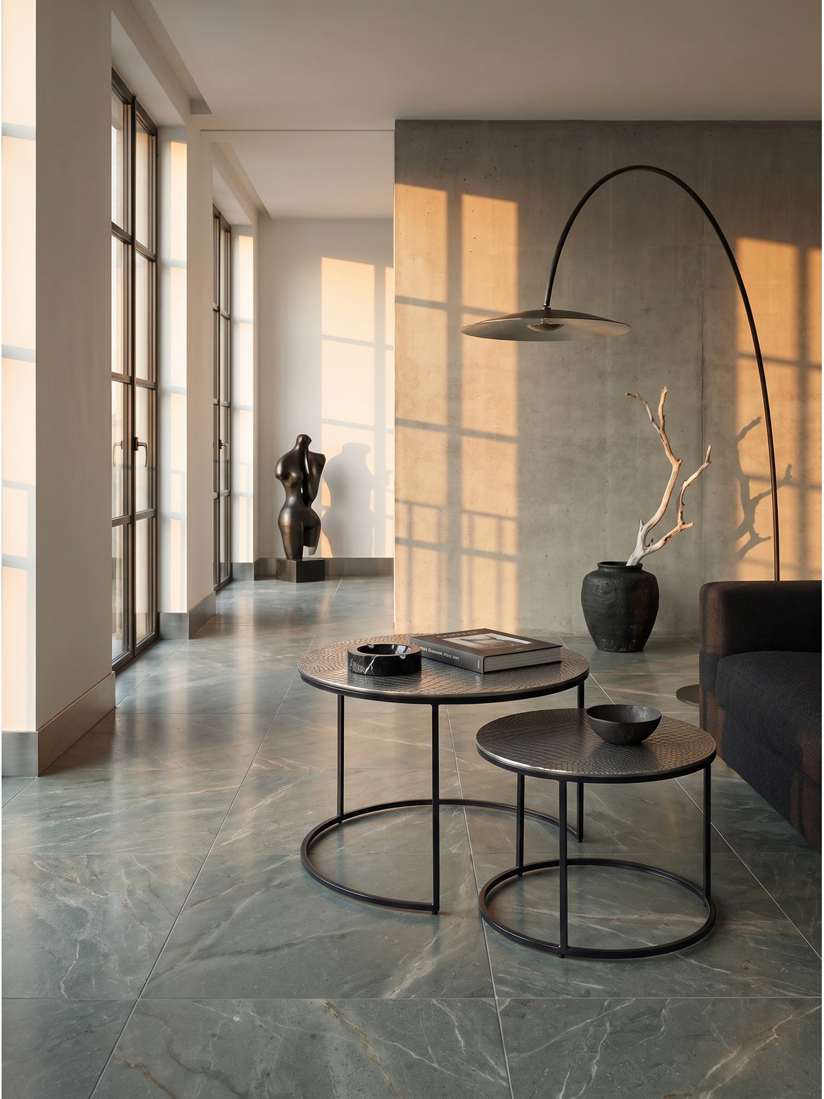 Zweiteiliger Aluminium-Couchtisch harmoniert mit einer schwarzen minimalistischen Bogenleuchte vor einer rauen glatten Sichtbetonwand.