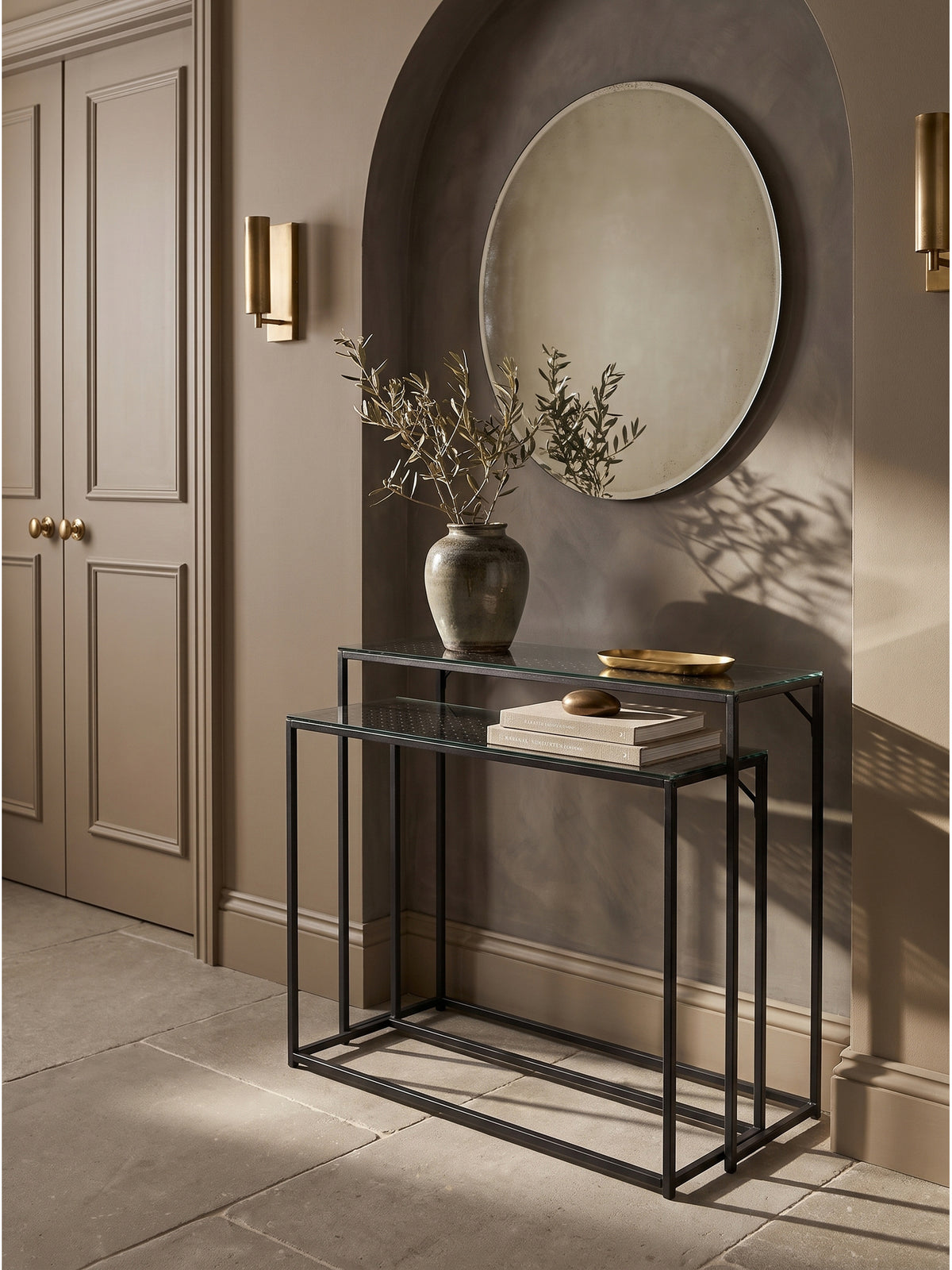 Schwarzes Konsolentisch Set aus Metall und Glas im eleganten Flur vor einer limewash Wand mit goldenen Wandleuchten_
