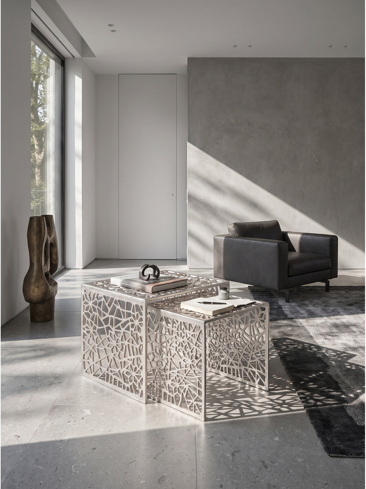 Silbernes Couchtisch-Set aus durchbrochenem Aluminium im minimalistischen Wohnzimmer vor einer hellgrauen Microzement-Wand auf anthrazitfarbenem Seidenteppich