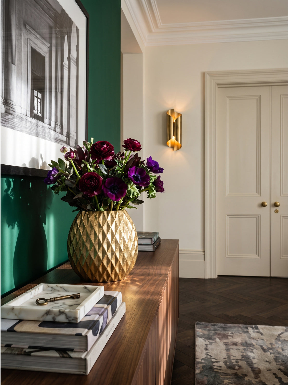 Schmale goldene Tischvase aus Aluminium arrangiert mit dunkelroten Trockenzweigen auf einem antiken Sideboard unter warm leuchtenden Messing-Wandleuchten._