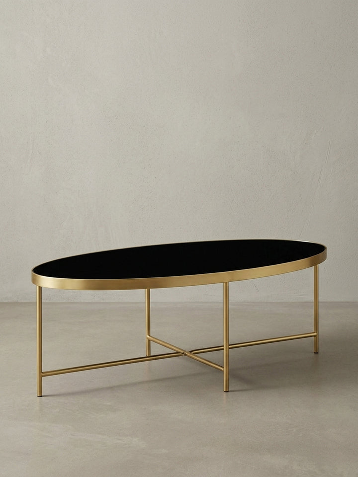 Moderner Lounge Tisch in ovaler Form aus der Dreiviertelperspektive, das gekreuzte Untergestell aus mattgoldenem Stahl ist unter der Glasplatte gut erkennbar._