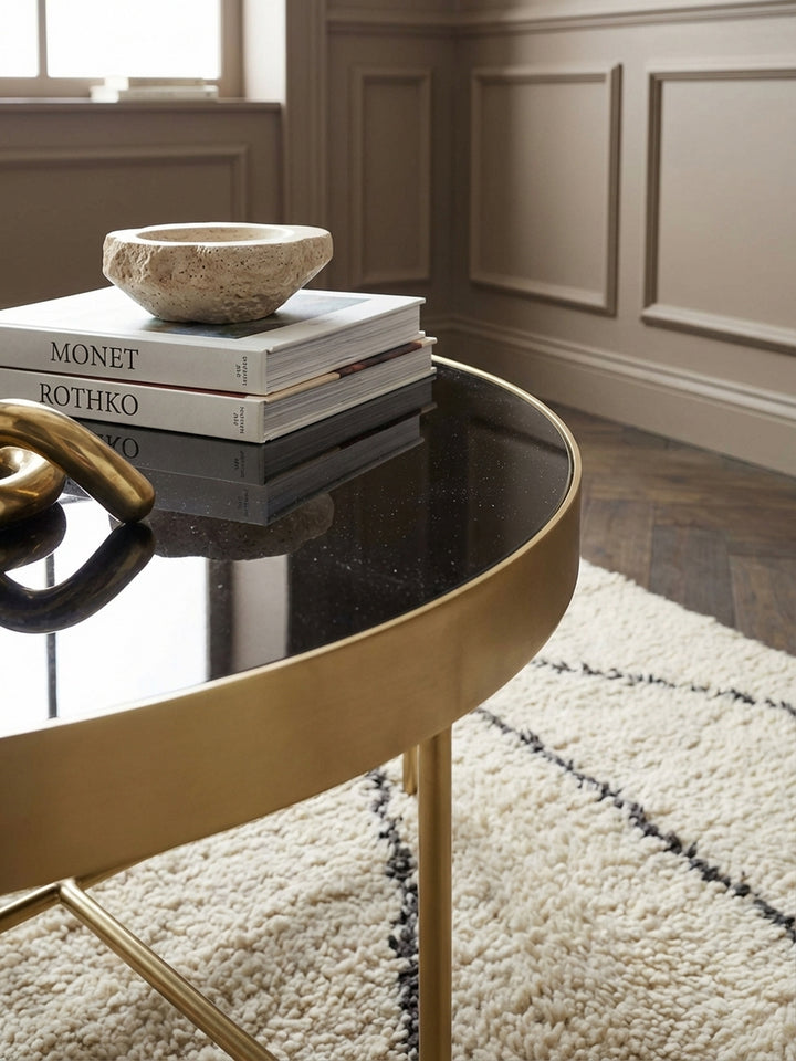 Nahaufnahme der Tischkante des ovalen Couchtisches, die den matten Goldton des Stahlrahmens und die spiegelglatte schwarze Glasplatte im Detail zeigt._