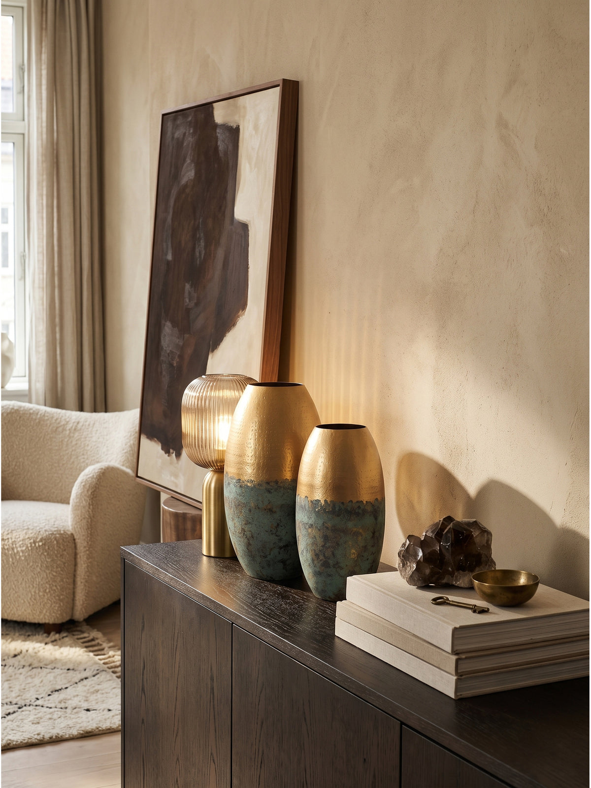 Elegante Tischvase aus massivem Aluminium mit goldenem Finish und einem getrockneten Treibholz-Ast arrangiert neben drei Leinen-Bildbänden auf dunklem Holz._