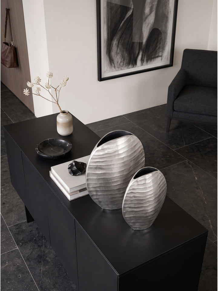 Elegantes Tischarrangement von oben betrachtet mit zwei glänzenden Metallvasen neben einem Fotografie-Bildband auf dunklem Stein._