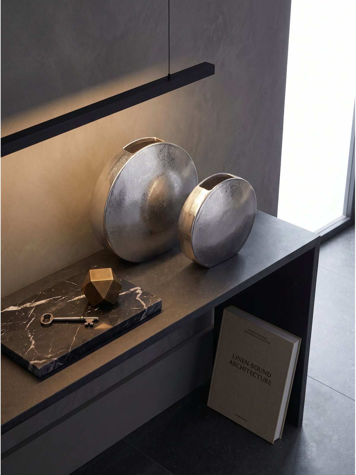 Zweiteiliges Vasen-Set aus strukturiertem Metall von oben auf einem Stein-Sideboard neben einem Bildband._