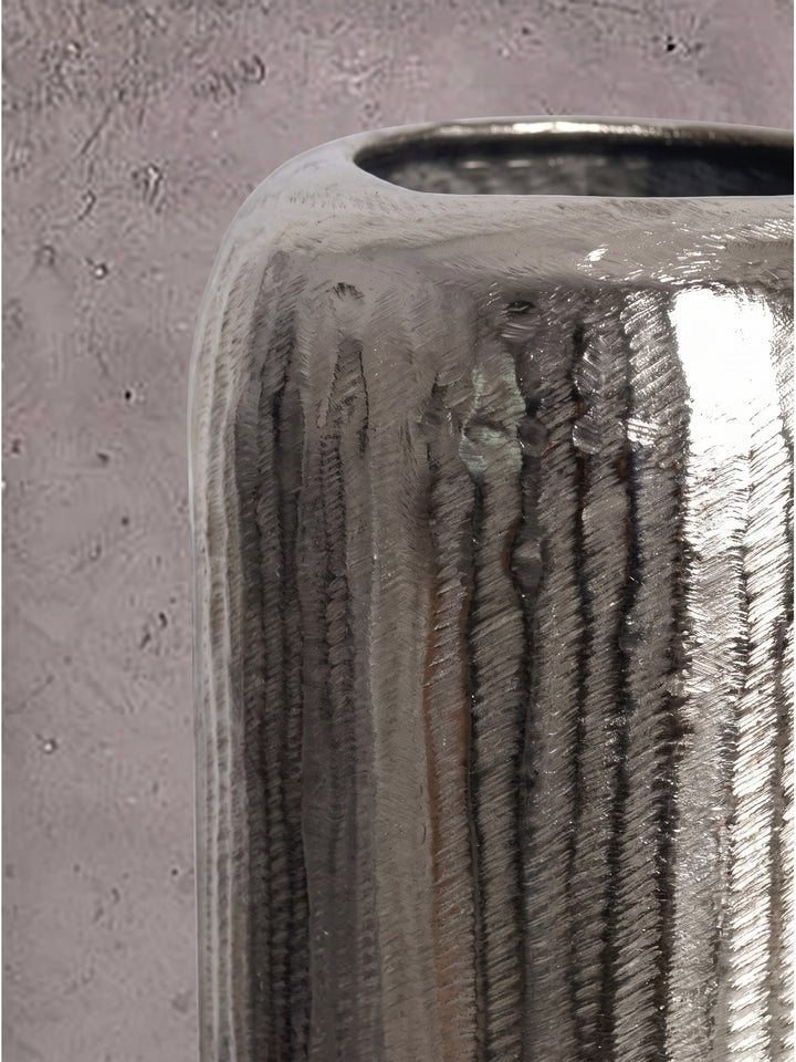 Nahaufnahme der stark geriffelten Aluminiumoberfläche der silbernen Dekovasen im Kontrast zur matten Steinwand._