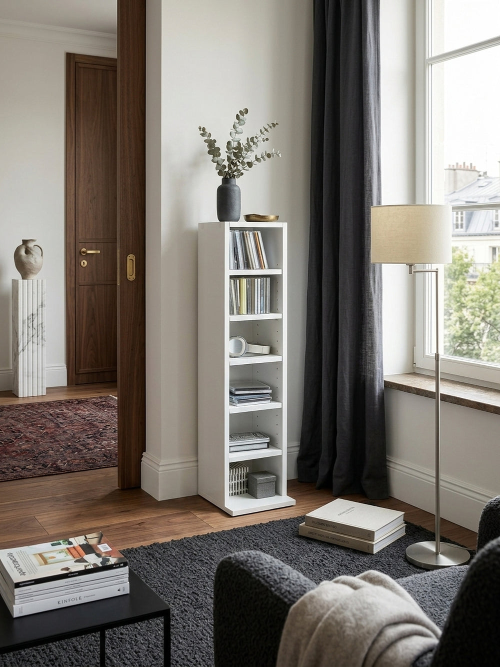 Schmales weißes Standregal mit 6 Fächern in eleganter Wohnzimmer-Lesecke auf dunklem Bouclé-Teppich, dekoriert mit Kunstbänden und Messing-Details._