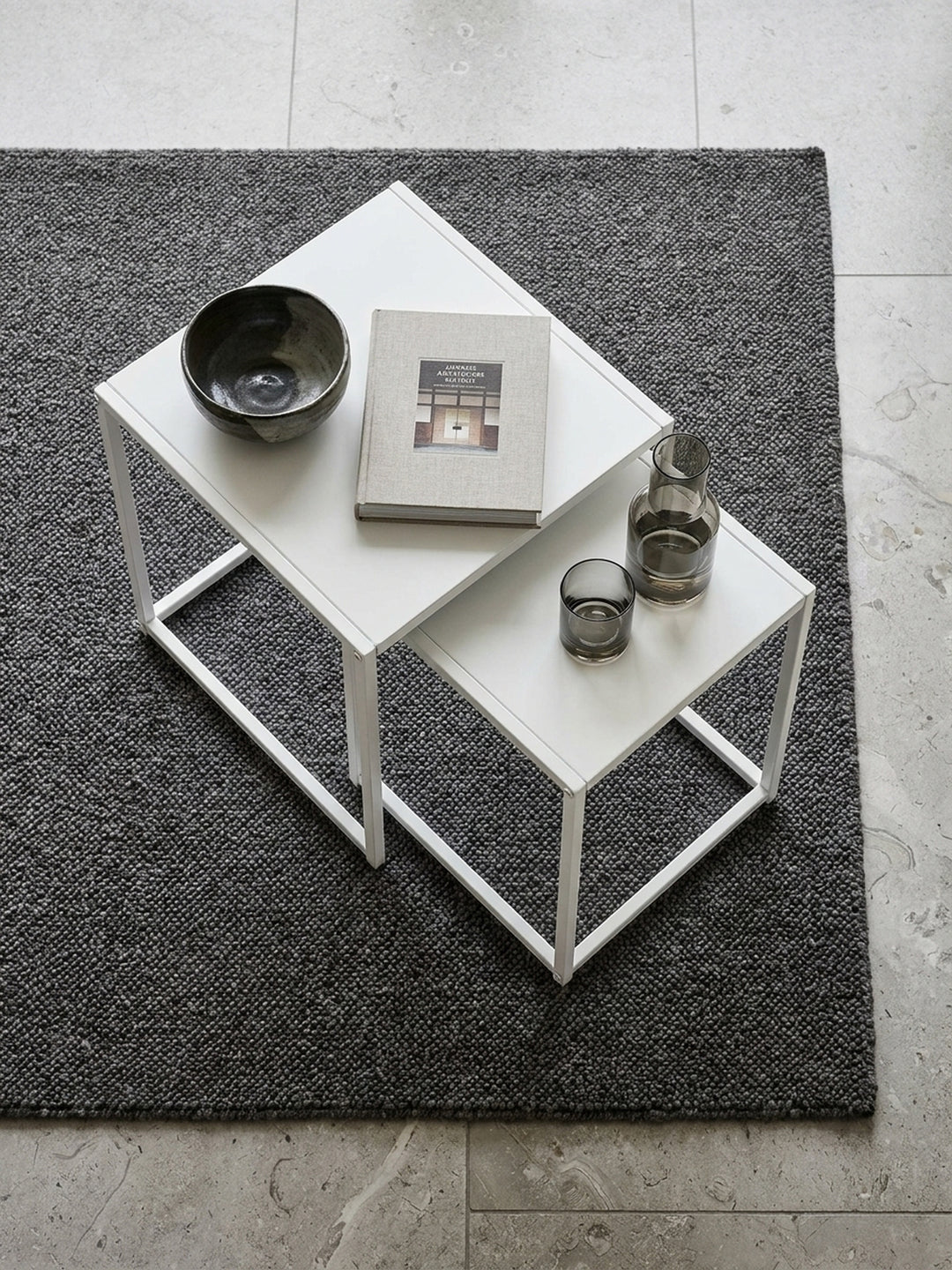 Aufsicht auf ein zweiteiliges Satztisch-Set aus weißem Stahl mit Teekanne auf der Ablage, arrangiert vor einem modernen Sofa auf großformatigen Kalksteinfliesen._