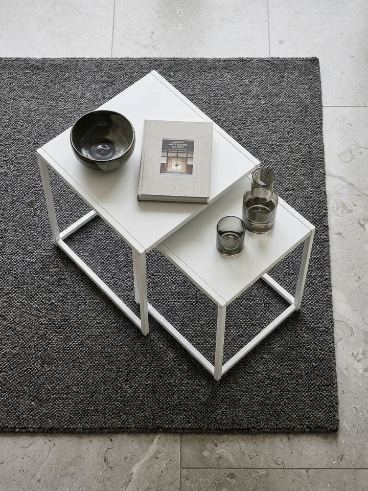Aufsicht auf ein zweiteiliges Satztisch-Set aus weißem Stahl mit Teekanne auf der Ablage, arrangiert vor einem modernen Sofa auf großformatigen Kalksteinfliesen._